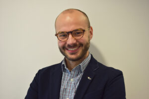 Smiling man with glasses in a blazer and checkered shirt, standing against a plain background.