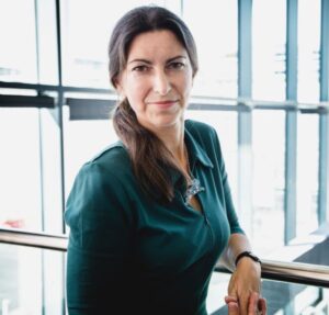 Woman in green top leaning on a railing, with large windows in the background, smiling.