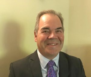 Smiling man in a suit with a patterned tie sitting in a room with beige walls.