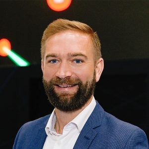 Smiling man with a beard in a blue blazer and white shirt, standing indoors.