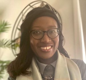 A person in glasses, wearing a scarf, smiles warmly indoors with a green plant in the background.