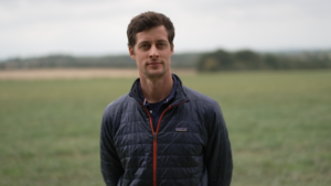 A person wearing a blue jacket stands outside in a field on a cloudy day.