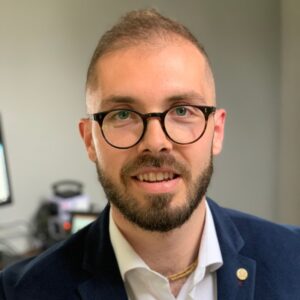 Man with glasses and a beard in a blazer, smiling at the camera indoors.