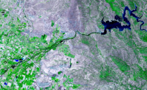 Satellite image of green farmland and rocky terrain with a winding river and a dark blue lake in the upper right.