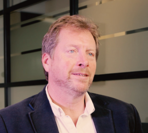 A man with a beard wearing a suit jacket and white shirt, sitting in front of a window with frosted glass.