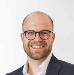 Man with glasses and beard smiling, wearing a dark suit jacket and white shirt against a plain background.
