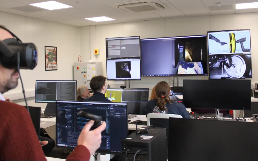 People in a control room work at computers while one person uses a VR headset and controller.
