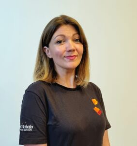 Person in a black shirt with short hair stands against a plain background, smiling slightly.