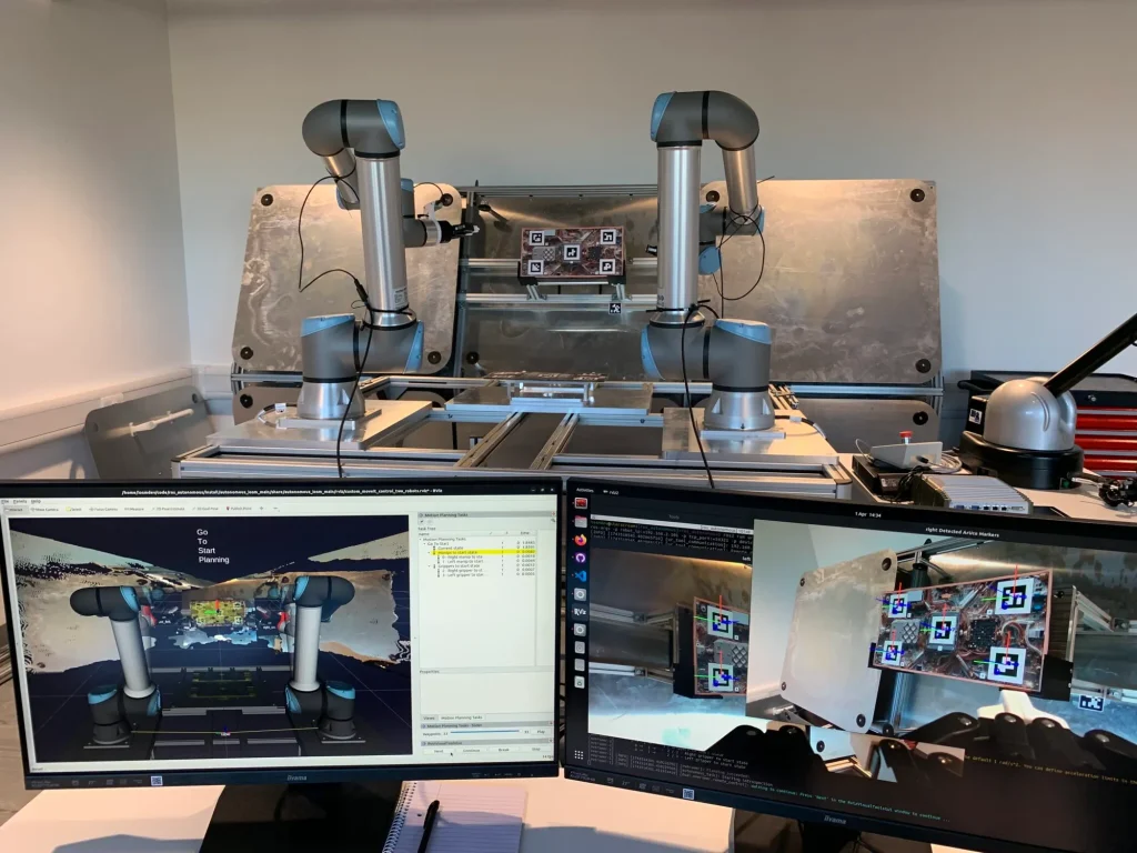 Two robot arms operate on a metal table; two monitors display robotics software and camera feeds.