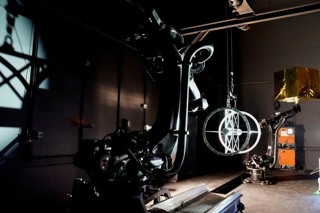 Two robotic arms operate in a dark room; one lifts a metallic spherical object with a geometric design.