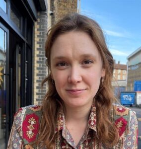 A person with long brown hair wearing a patterned shirt sits outside near brick buildings, looking at the camera.