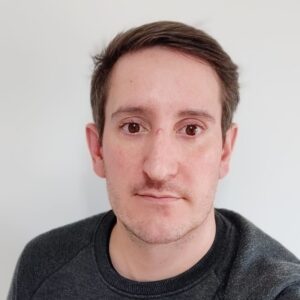 Person with short dark hair and a neutral expression wearing a dark sweater against a plain light background.