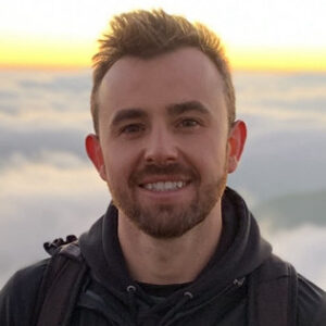 A man with short hair and a beard smiles, standing outdoors with a cloudy sky and sunrise or sunset in the background.