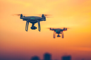 Two drones flying against an orange sunset sky, with blurred city buildings in the background.