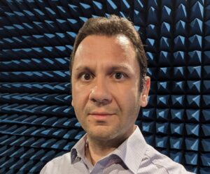 Man in a striped shirt in an anechoic chamber with blue, foam pyramidal panels.