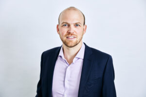 A man with a beard wearing a navy suit and light purple shirt, standing against a plain white background.