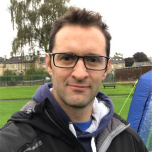 Man with glasses and short hair in an outdoor setting with grass and buildings in the background.