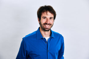 Smiling man with dark hair and beard wearing a blue shirt stands against a plain white background.