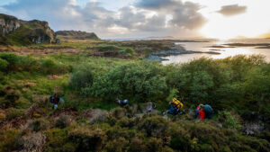 Hikers with backpacks traverse lush terrain near a rocky coastline at sunset, with hills and water in the distance.
