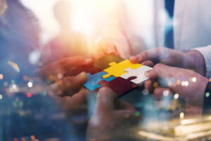 Hands of several people holding interlocking puzzle pieces against a blurred, colorful background.