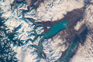 Satellite view of a turquoise lake surrounded by snowy mountains and brown terrain.
