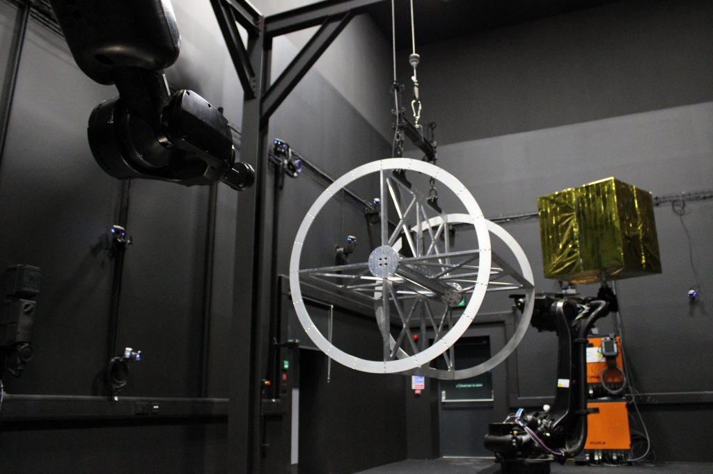 A robotic arm holds a large metal ring structure in a dark industrial room with various equipment.