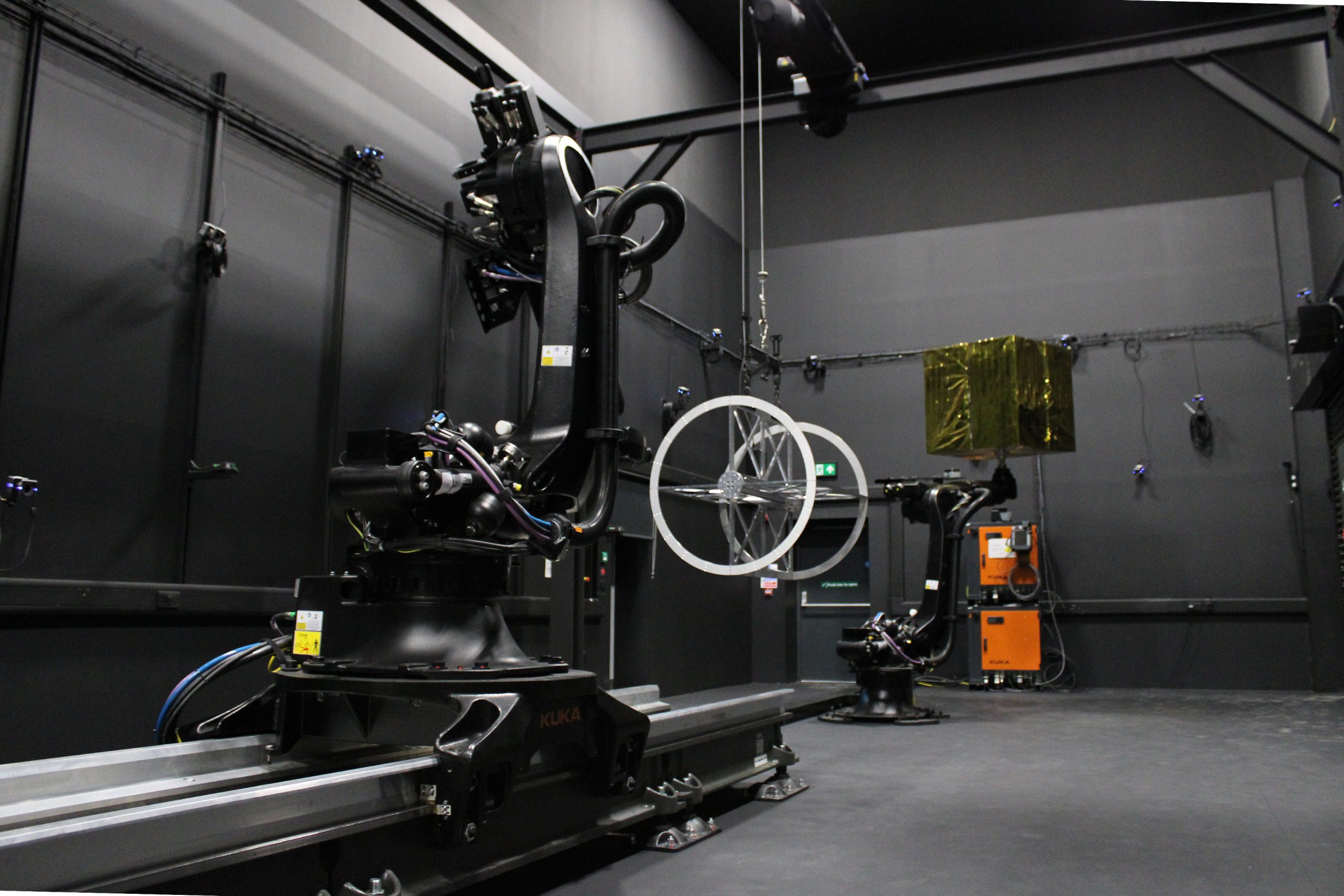 Two industrial robotic arms operate in a dark lab; one holds a circular metal frame suspended in the air.
