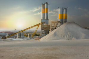 Industrial site with large gravel pile, conveyor belt, and towers under a cloudy sky at sunset.