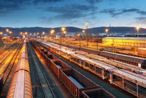Railway yard at dusk with lit overhead lights, multiple train tracks, and freight trains against a hilly backdrop.