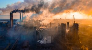 Aerial view of an industrial area emitting thick smoke from stacks, set against a colorful, hazy sunset sky.