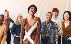 A diverse group of seven women standing together, smiling and laughing in front of a light-colored background.