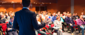 Person in a suit giving a presentation to a seated audience in a conference room.