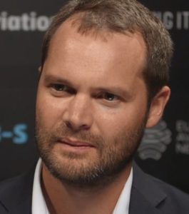 Man with short dark hair and beard, wearing a suit, against a dark background with text.