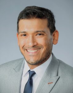 A smiling man in a gray suit and black tie against a light blue background.
