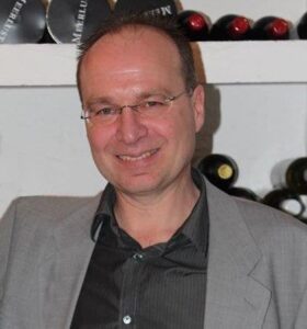 Person wearing glasses and a gray suit jacket, standing in front of shelves with wine bottles.