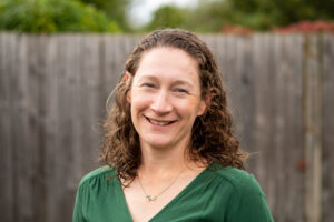 Smiling woman with curly hair stands outdoors in front of a wooden fence, wearing a green top.