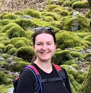 Person smiling in front of lush green moss-covered rocks, wearing a black top with sunglasses on their head.
