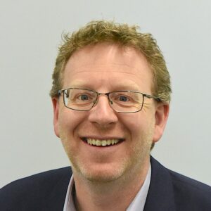 Smiling man with glasses and wavy hair wearing a dark blazer and light shirt in front of a plain background.
