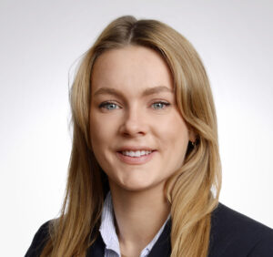 Person with long blonde hair smiling, wearing a dark blazer and striped shirt, against a light gray background.