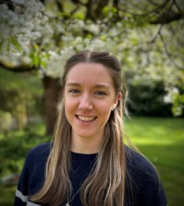A woman smiling in a garden with blooming trees and a blurred green background.