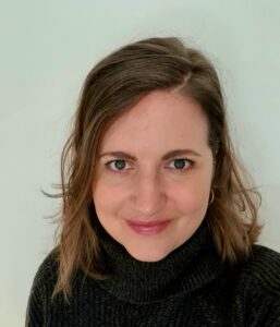 Woman with shoulder-length brown hair and hoop earrings, smiling at the camera, wearing a black turtleneck sweater.