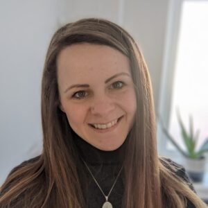 Person with long brown hair smiling, wearing a black top and necklace, with a blurred plant in the background.