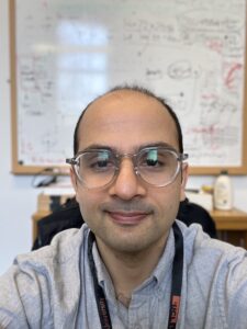 Person wearing glasses and a gray shirt, sitting in front of a whiteboard with writing and diagrams.