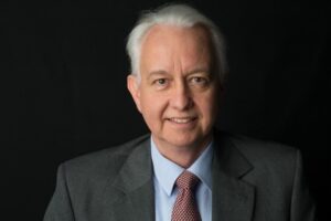 Smiling man with white hair in a gray suit and red tie against a black background.
