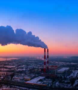 Factory with white and red chimneys emitting smoke against a colorful sunset sky over an industrial landscape.