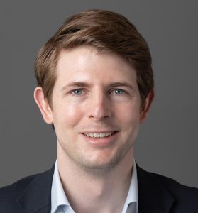 A person with short brown hair smiles, wearing a light blue shirt and dark blazer, against a plain gray background.