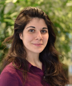 Woman with long dark hair smiling, wearing a purple shirt, standing in front of blurred green foliage.
