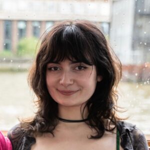 Person with dark hair and a choker smiling indoors with a blurred building in the background.