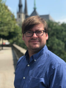 Man in a blue checkered shirt and glasses stands outdoors with trees and a building in the background.