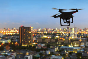 A drone hovers over a cityscape with blurred, glowing lights at dusk.